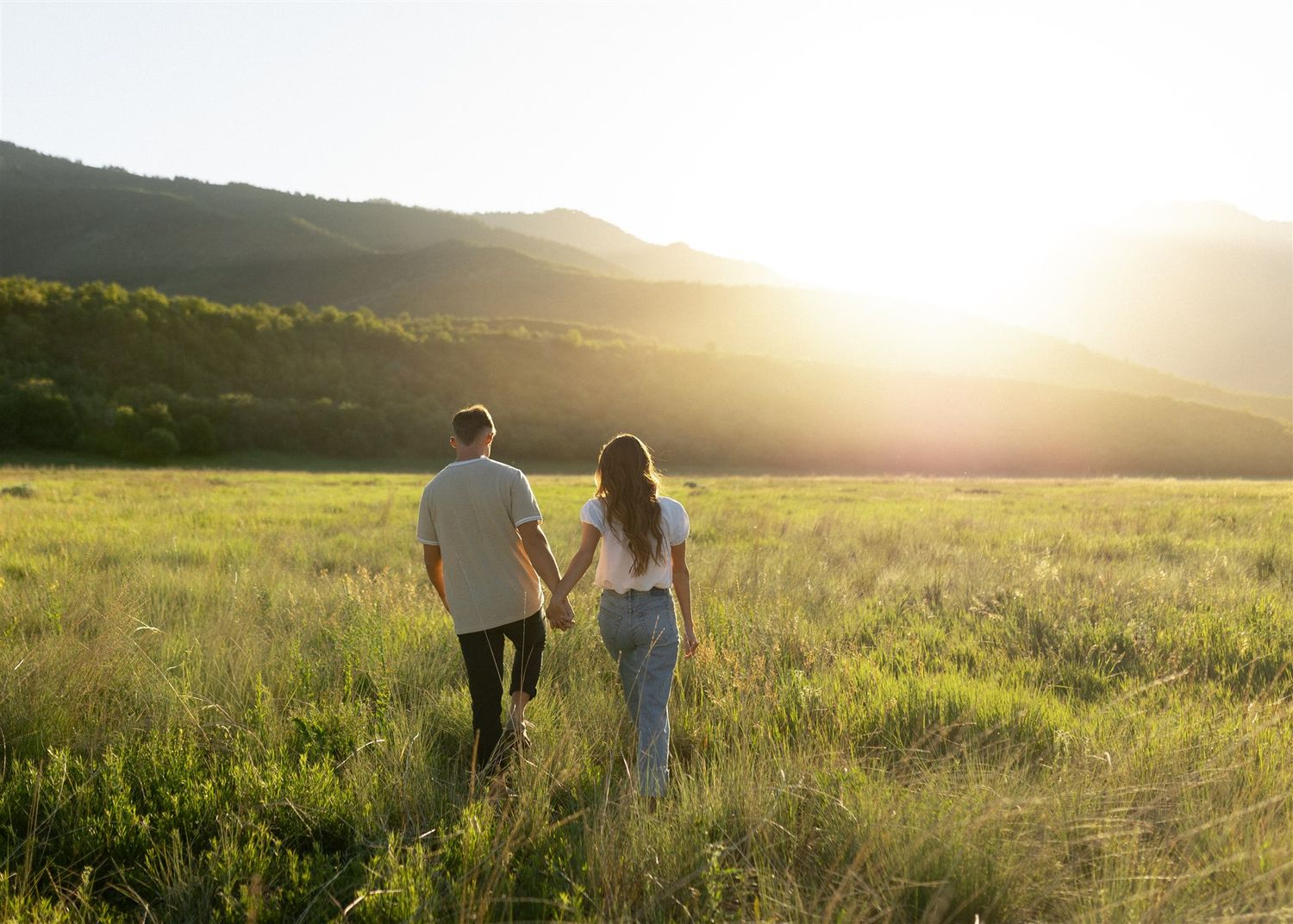 Couple holding hands walking through a sunlit green meadow with mountains in the golden hour light.