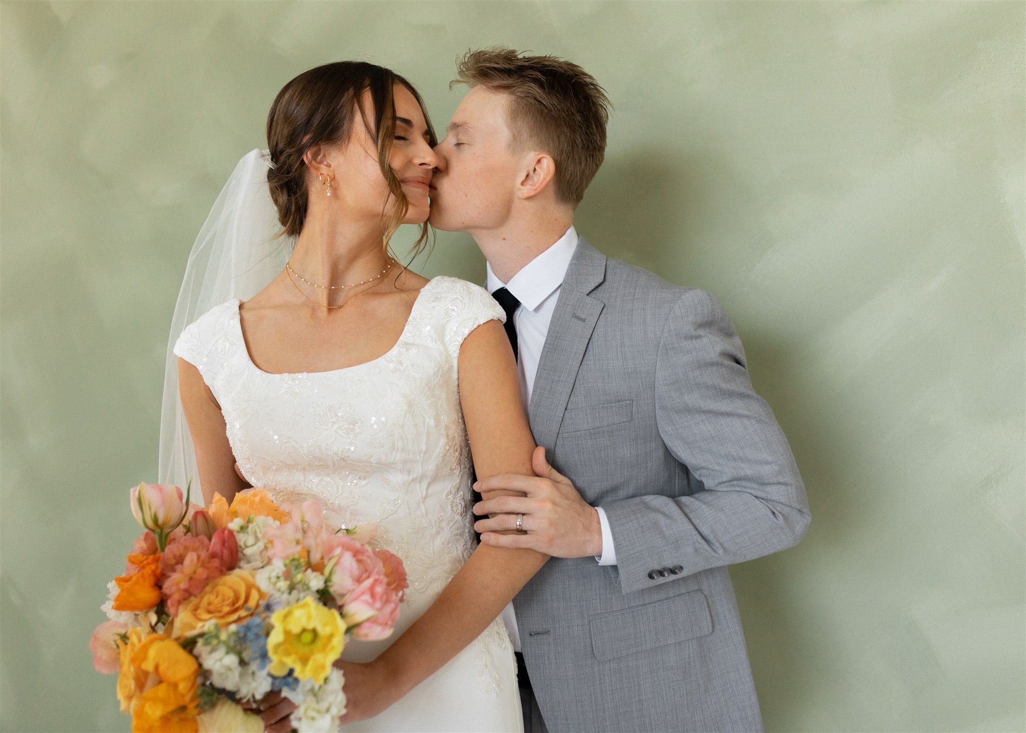 Bride and groom kissing, she holds colorful bouquet, he wears gray suit, green backdrop.