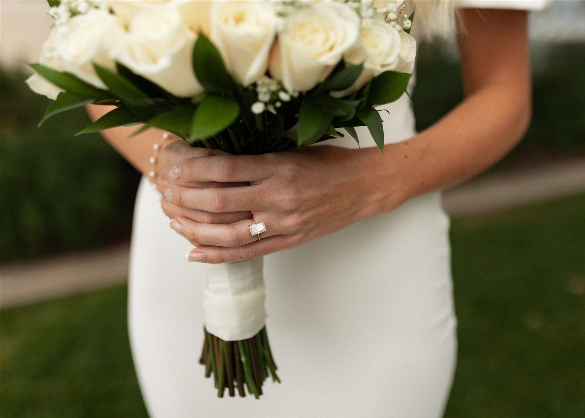 A bride in a white dress holding an elegant bouquet of white roses with green foliage.
