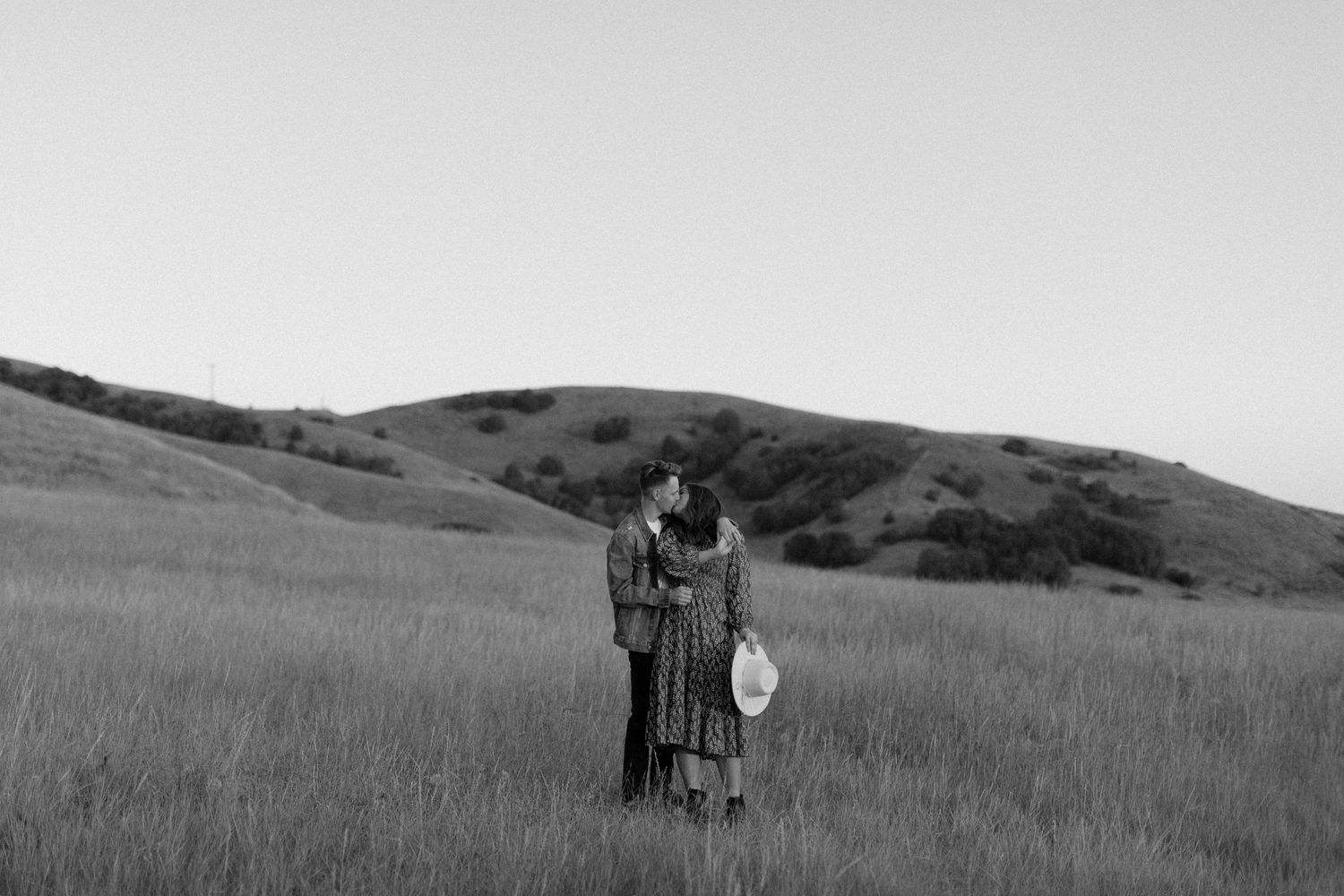 Black and white photo of a couple embracing in a grassy meadow with rolling hills in the background.
