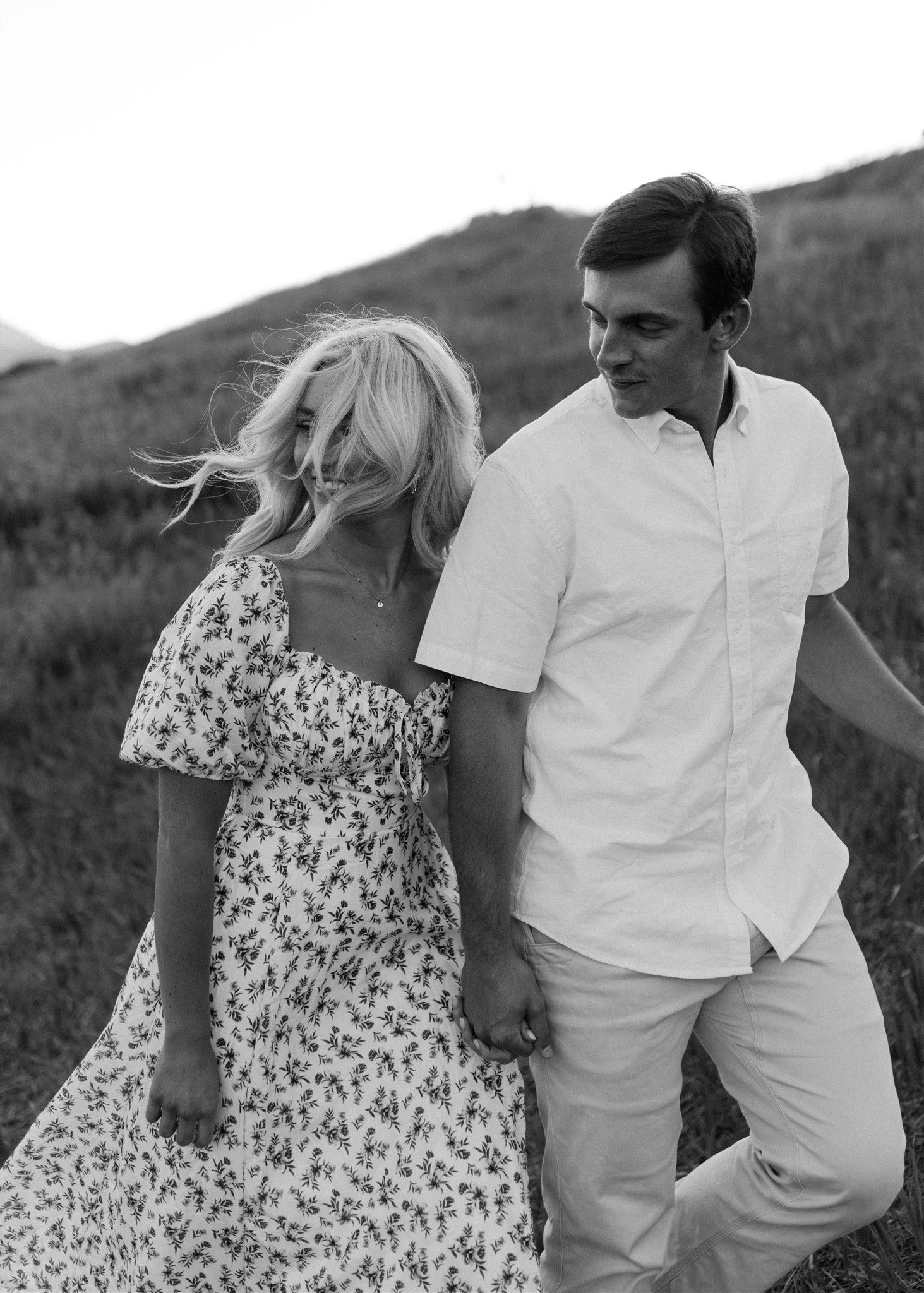 Black and white photo of blonde woman in floral dress and man in white shirt on hillside.