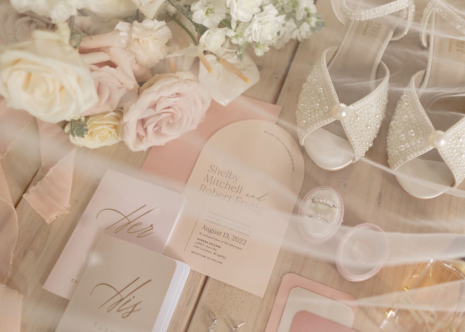 Elegant blush wedding stationery flat lay with roses, ribbon, and His and Hers vow books.